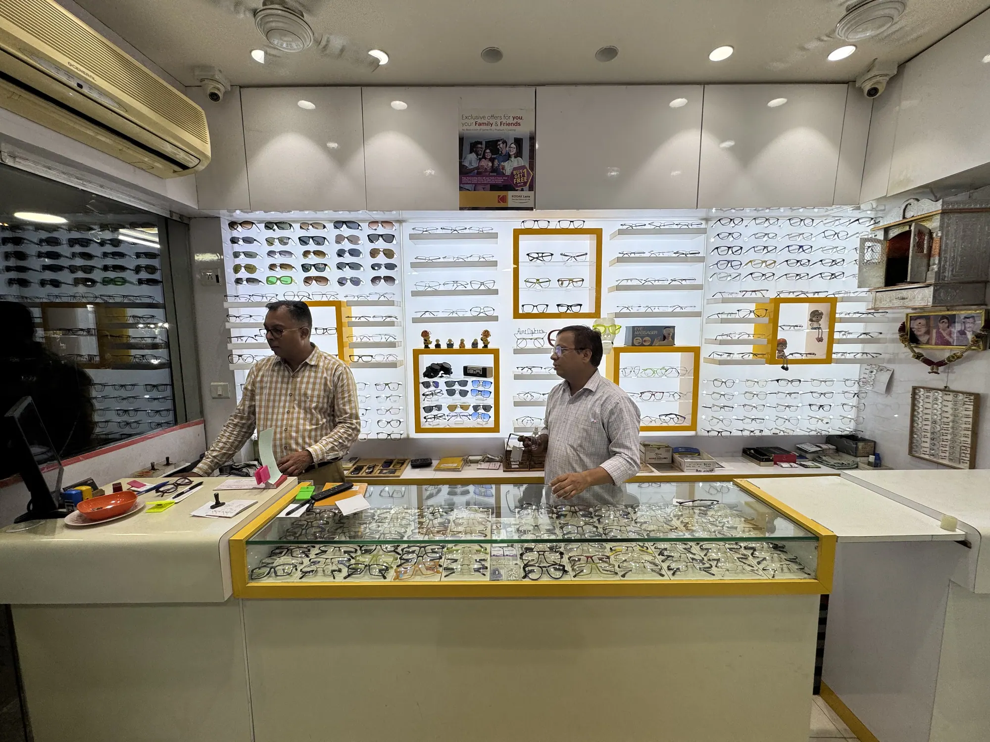 Inside Ami Optics at Jivdani Chowk, with the brothers at the counter and frames on every wall